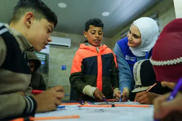 A humanitarian worker wearing a blue vest helps a group of children draw together around a table, while a boy in a bright orange jacket stands in the center of the activity.