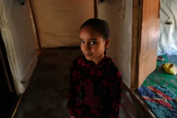 A young girl stands inside a temporary shelter, looking toward the camera, with fabric walls and a doorway behind her.