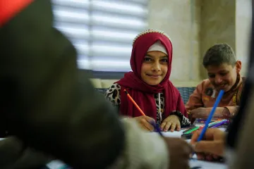 A girl wearing a burgundy headscarf smiles while drawing with colored pencils during a classroom activity.